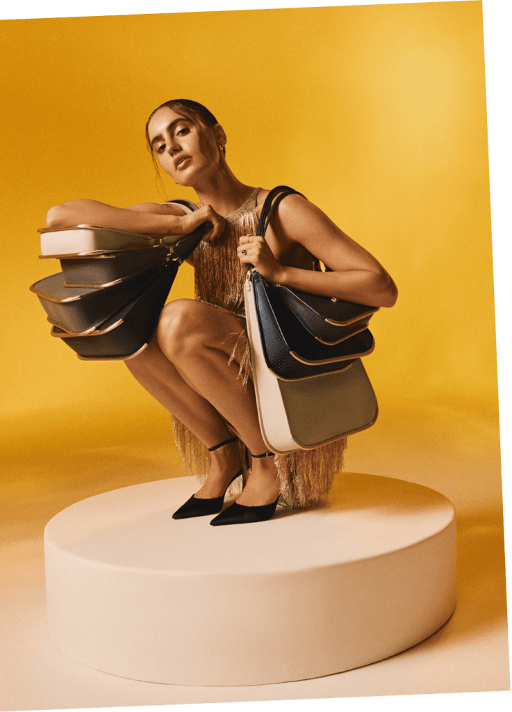 A woman in a gold dress sits on a pedestal, holding several VAULIE ICON luxury bags draped over her shoulder. The elegant bags are resting on her knees as she poses, highlighting their sophisticated design and luxurious design.