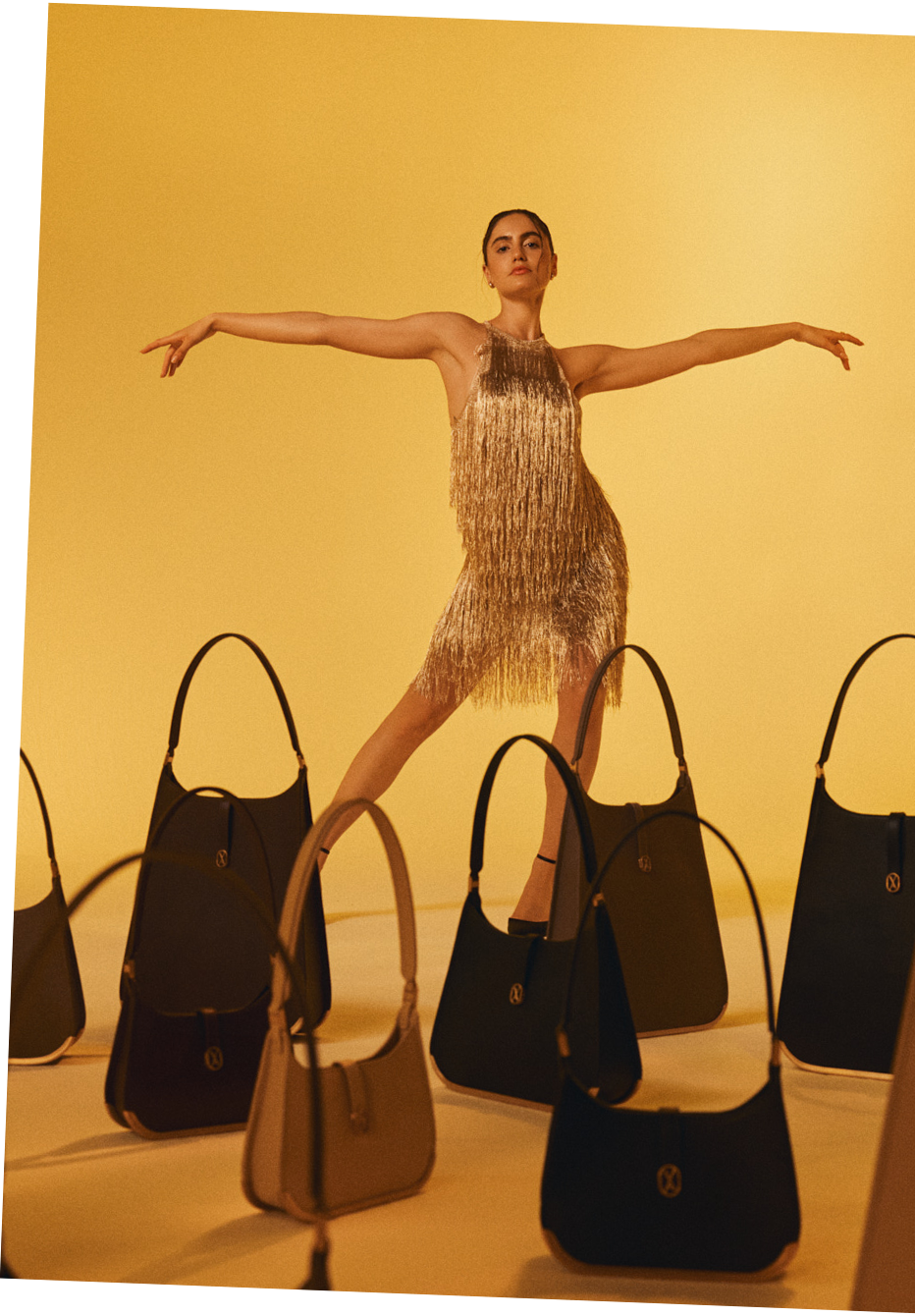 A woman in a gold dress stands proudly with her arms open, surrounded by a collection of luxury Vaulie bags arranged on the floor in front of her.