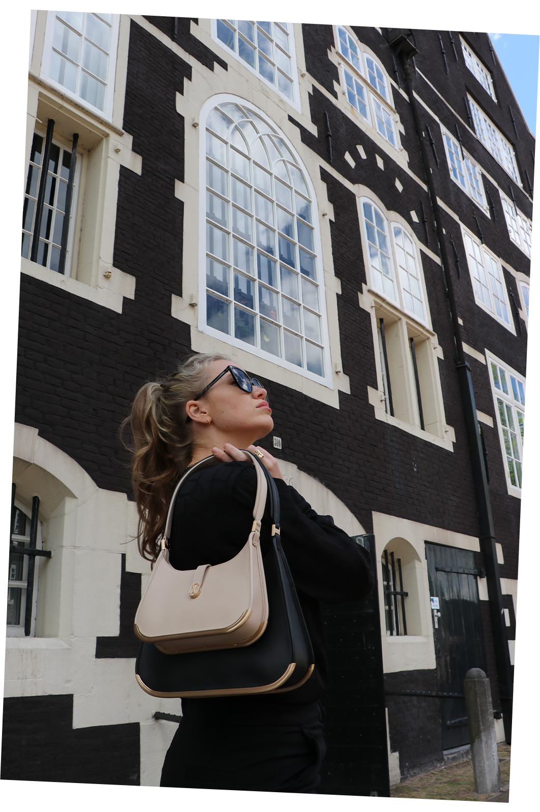 A proud woman with blond hair and large black sunglasses stands in front of a black-and-white building in Amsterdam. She holds two sizes of luxury bags over her shoulders in black and beige, both featuring a golden metal bottom.