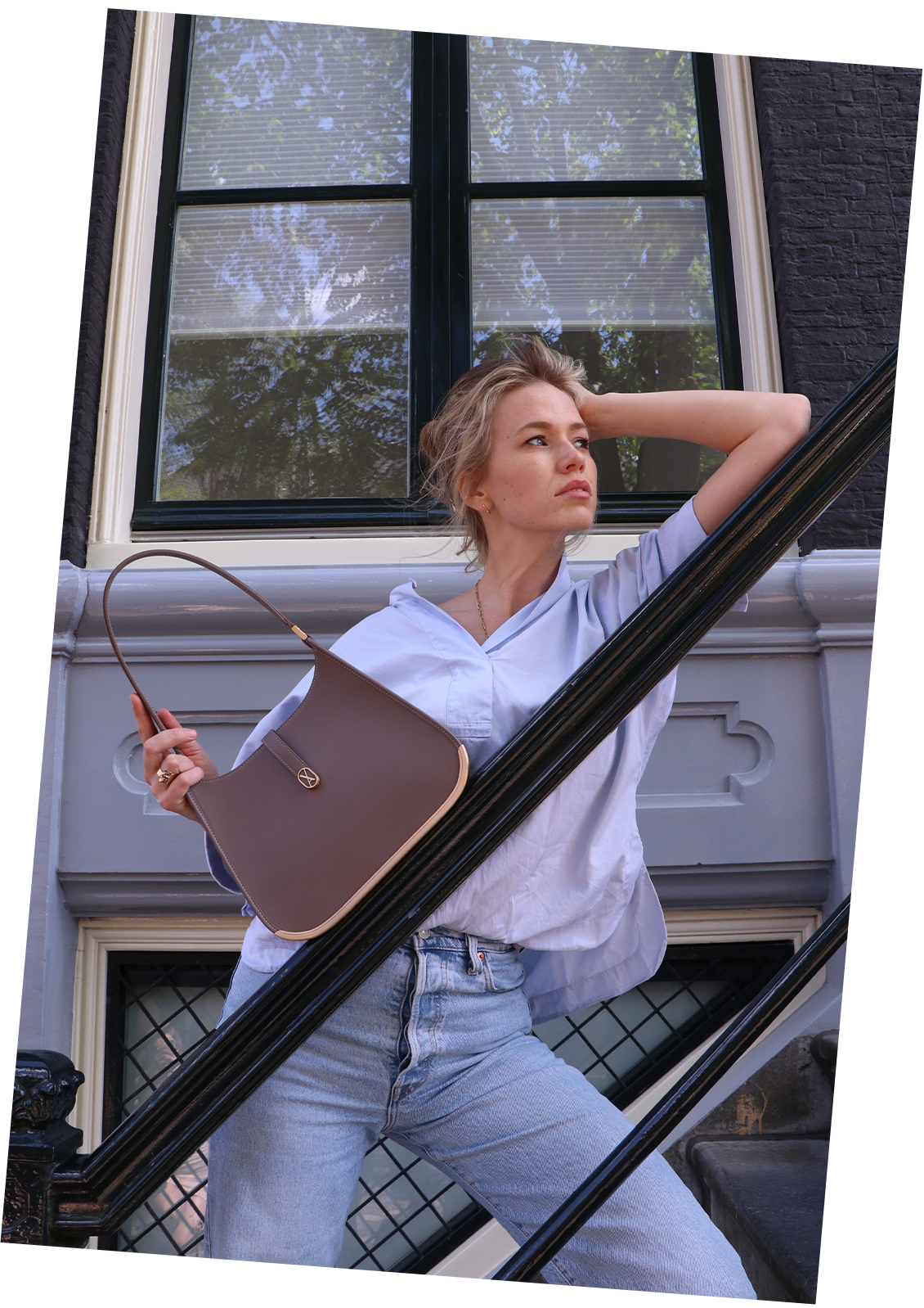 A woman leans on a stair in Amsterdam, resting her head on one hand while holding a taupe luxury Vaulie bag with a golden bottom in the other. The bag rests on the stair.