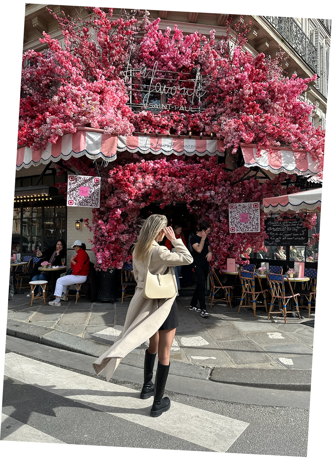 A chic woman stands in front of La Favorite Saint Paul in Paris, with her beige coat and blond hair flowing in the wind. A beige luxury Vaulie bag with a golden bottom hangs on her shoulder.