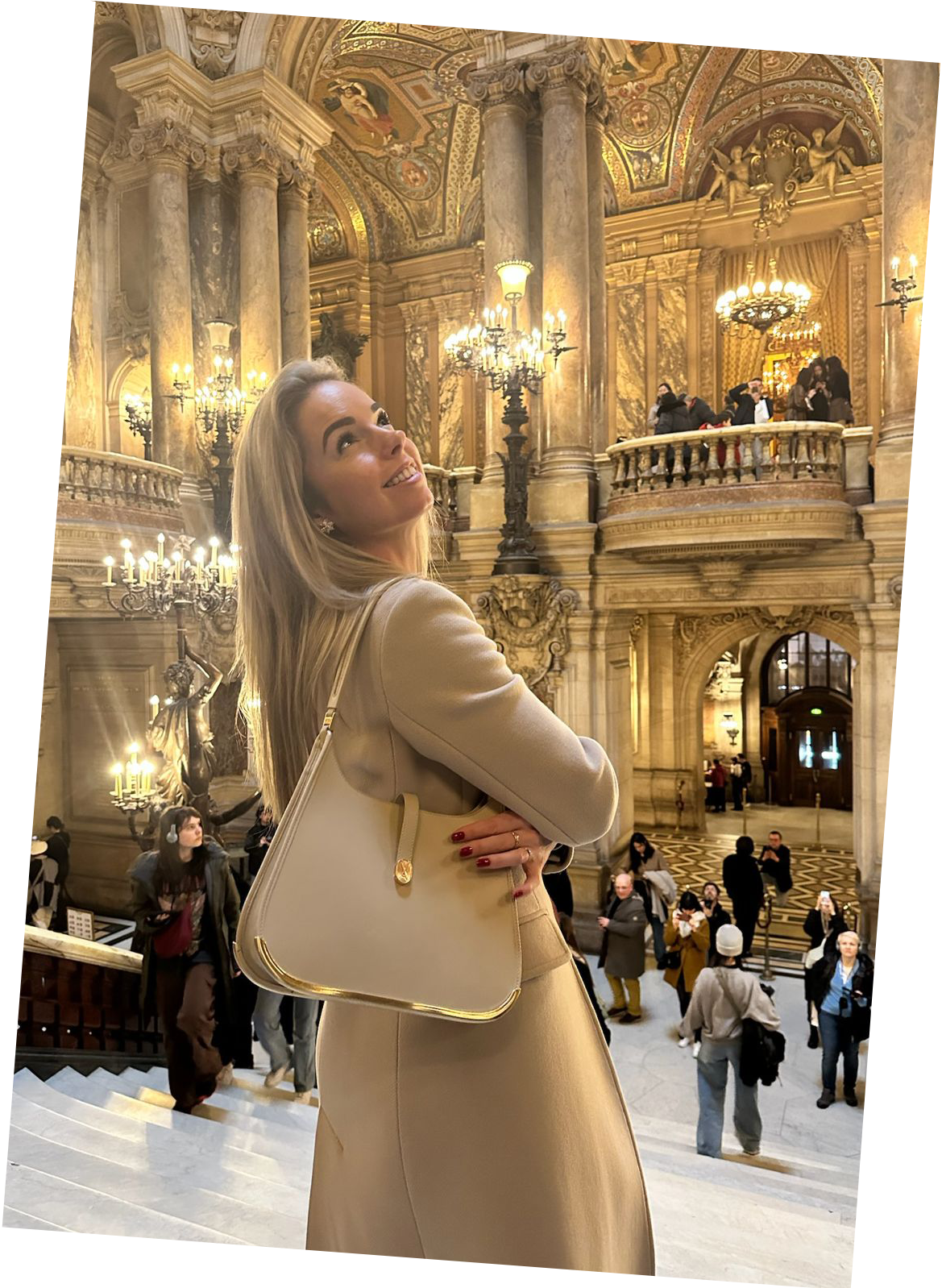 A classy woman inside the opera in Paris gazes upward, admiring the architectural ambiance. She carries a beige luxury Vaulie bag with a golden bottom on her shoulder.