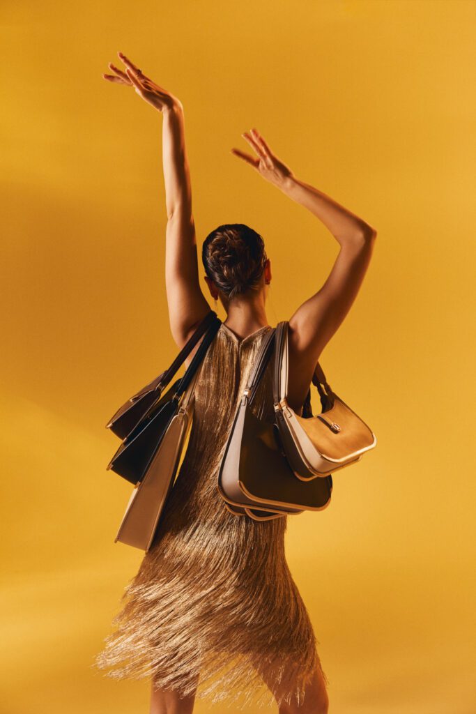 A woman in a gold fringed dress dances with her hands in the air, with several luxury Vaulie bags draped over her shoulders. The ICON bags feature various leather colors and an elegant golden bottom.