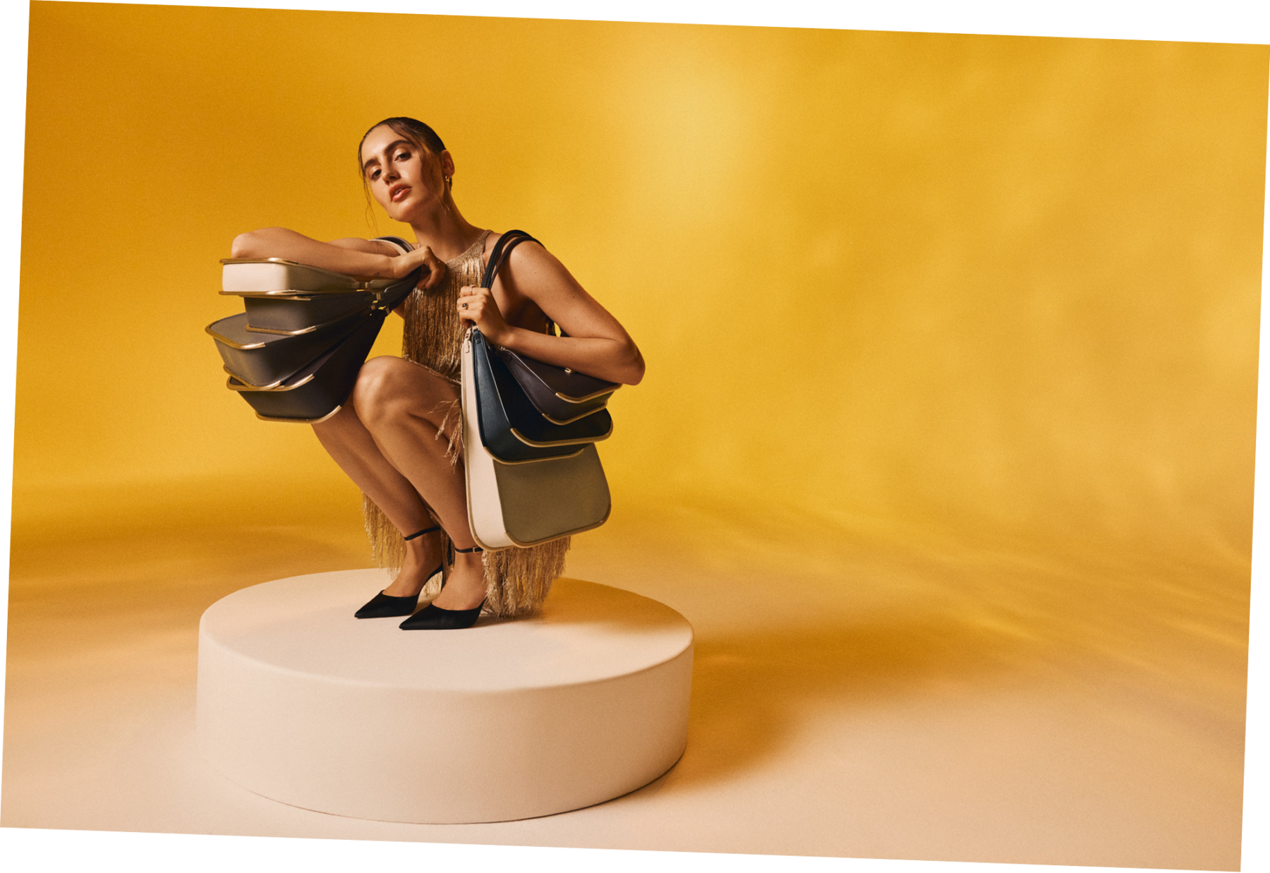 A woman in a gold dress sits on a pedestal, holding several VAULIE ICON luxury bags draped over her shoulder. The elegant bags are resting on her knees as she poses, highlighting their sophisticated design and luxurious design.