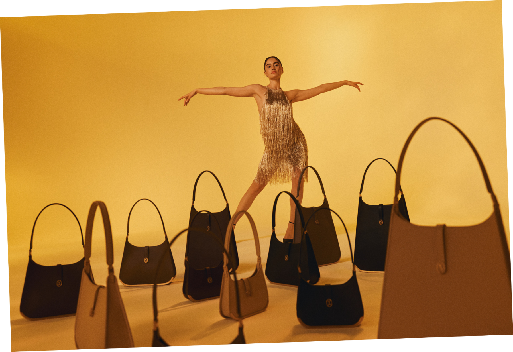 A woman in a gold dress stands proudly with her arms open, surrounded by a collection of luxury Vaulie bags arranged on the floor in front of her.