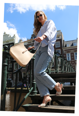 A chic woman walks down a bridge in Amsterdam, swinging a beige luxury bag towards the camera. The bag features a golden metal bottom and has feminine round curves.