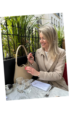 A luxury woman sits on a terrace in Paris, opening a bag placed on a marble table. The leather vaulie bag features a golden metal bottom.