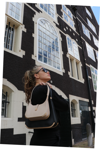 A proud woman with blond hair and large black sunglasses stands in front of a black-and-white building in Amsterdam. She holds two sizes of luxury bags over her shoulders in black and beige, both featuring a golden metal bottom.