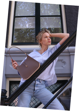 A woman leans on a stair in Amsterdam, resting her head on one hand while holding a taupe luxury Vaulie bag with a golden bottom in the other. The bag rests on the stair.