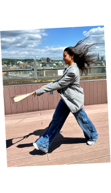 A businesswoman walks on a rooftop with her hair flowing in the wind, swinging a beige luxury Vaulie bag.