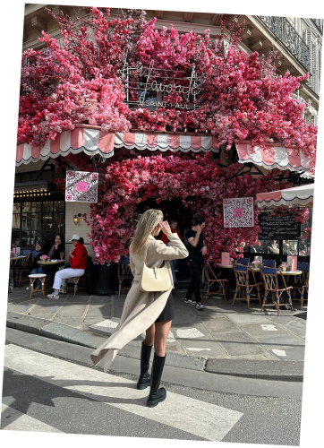 A chic woman stands in front of La Favorite Saint Paul in Paris, with her beige coat and blond hair flowing in the wind. A beige luxury Vaulie bag with a golden bottom hangs on her shoulder.