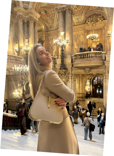 A classy woman inside the opera in Paris gazes upward, admiring the architectural ambiance. She carries a beige luxury Vaulie bag with a golden bottom on her shoulder.